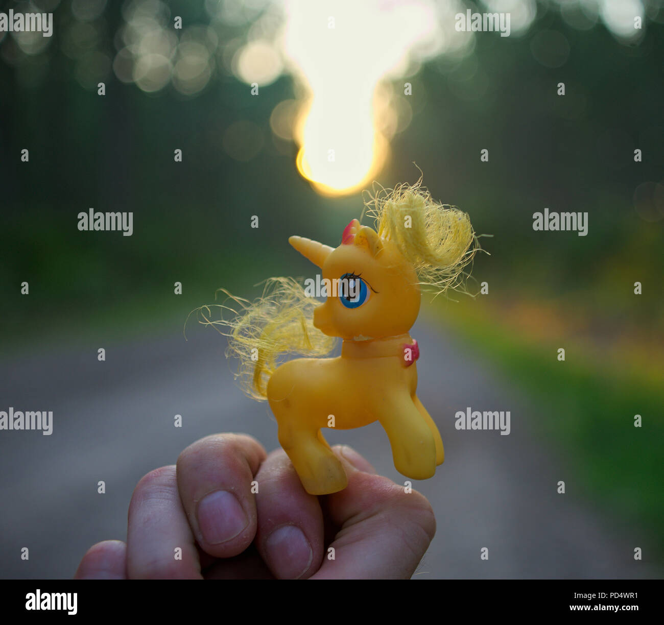 Hand holding yellow toy unicorn, forest road and sunset at background Stock Photo