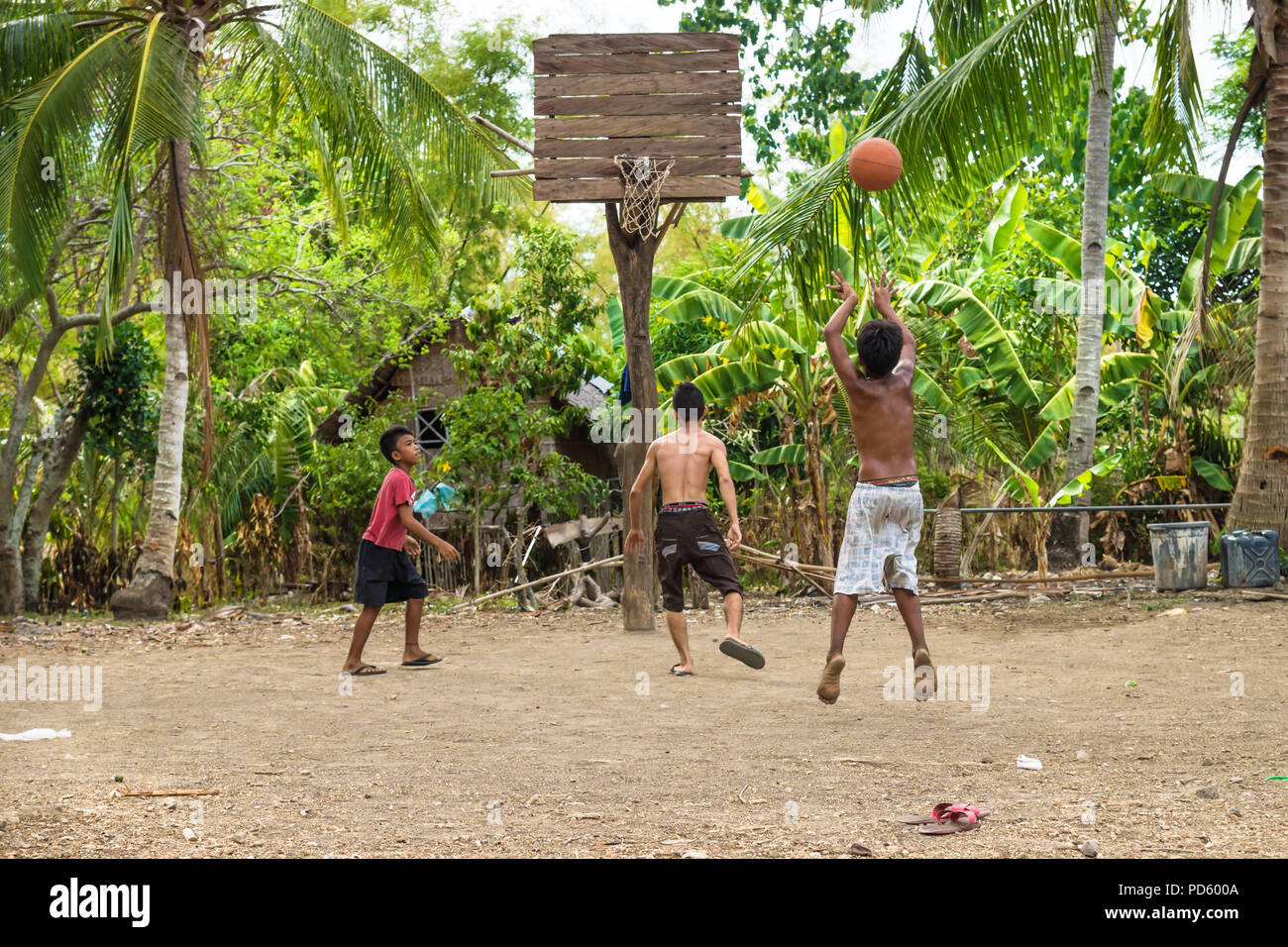 Sandugan, Siquijor, Philippines - May 13 2013: Three kids playing basketball with one kid jumping at sand field with old wooden basketball basket Stock Photo