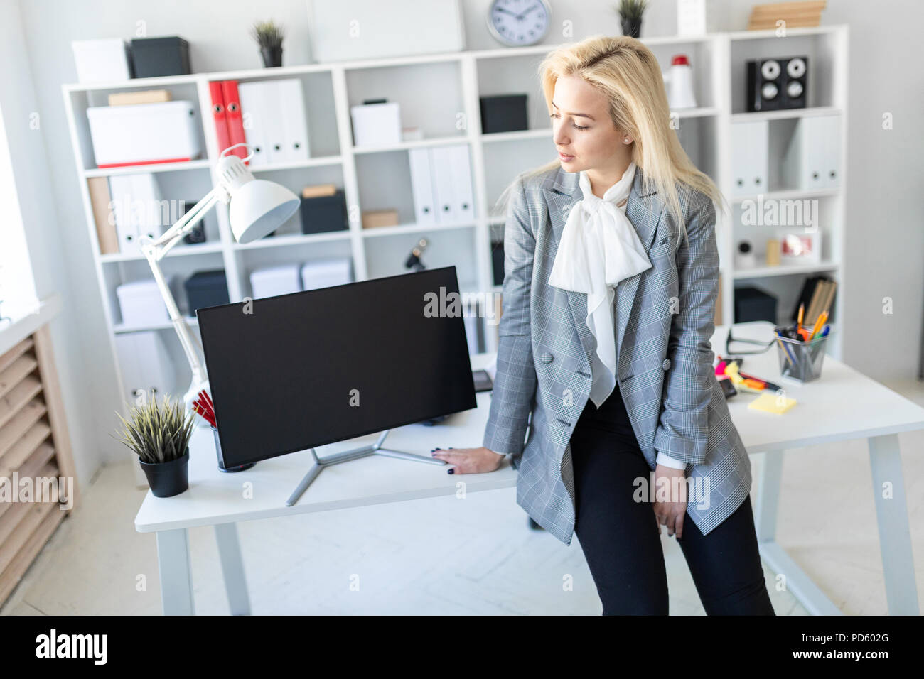Young girl stands in office, leaning on desk. Nearby is a monitor Stock