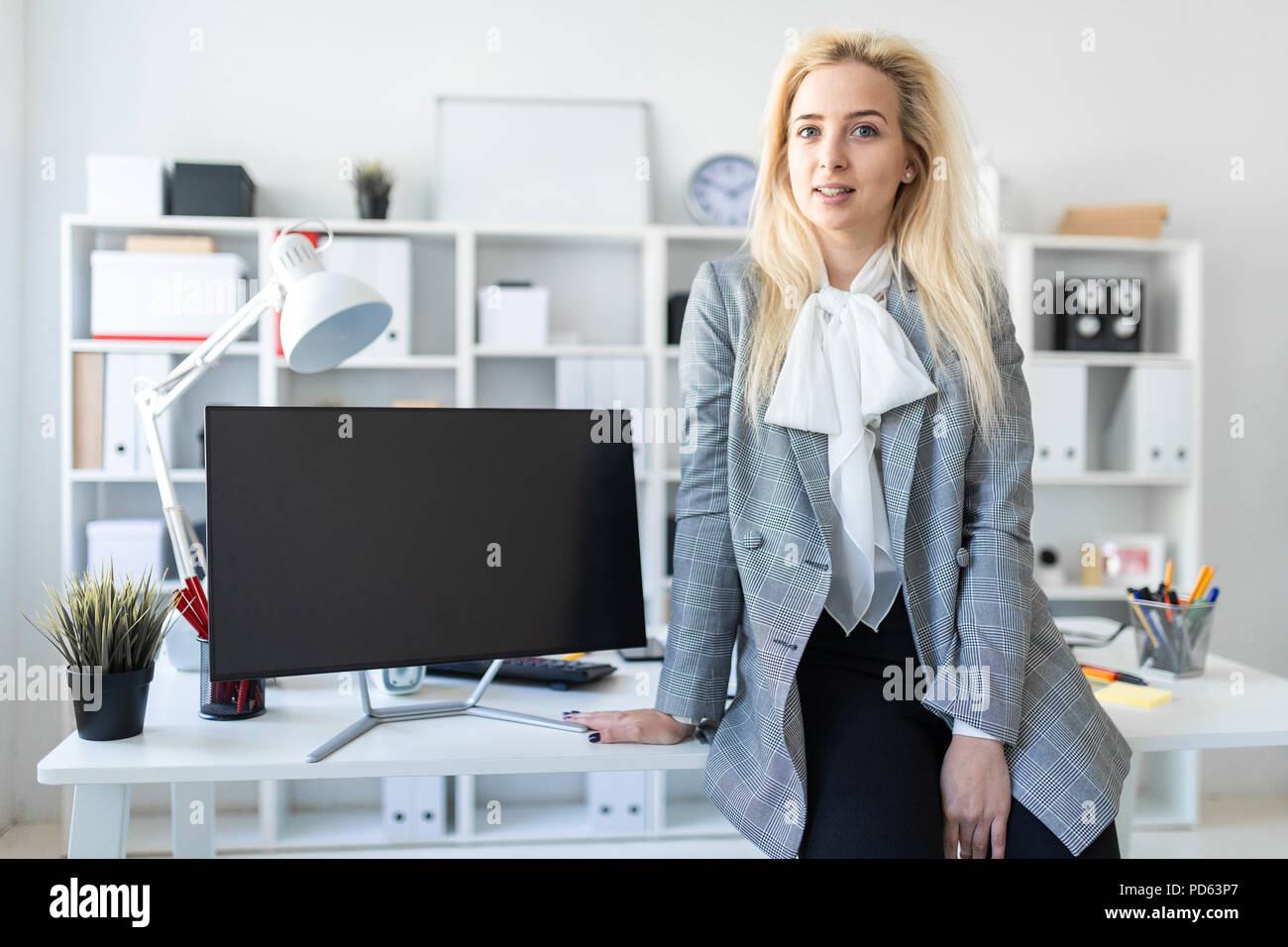 Young girl stands in office, leaning on desk. Nearby is a monitor Stock