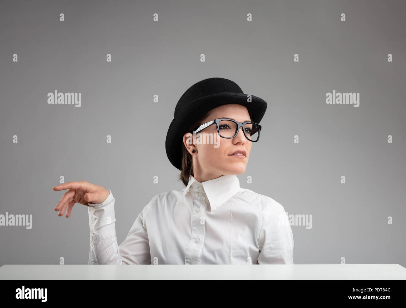 Elegant diva in a vintage bowler hat and glasses gesturing with her hand and looking off to the side with a superior proud expression against a grey b Stock Photo