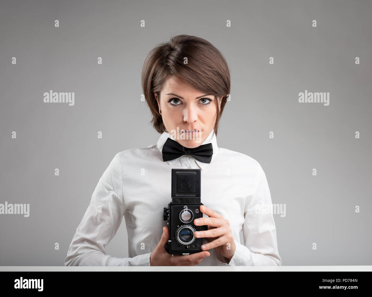 Stylish vintage female photographer wearing a bow tie holding her camera to her chest as she composes her image with a serious expression over grey Stock Photo