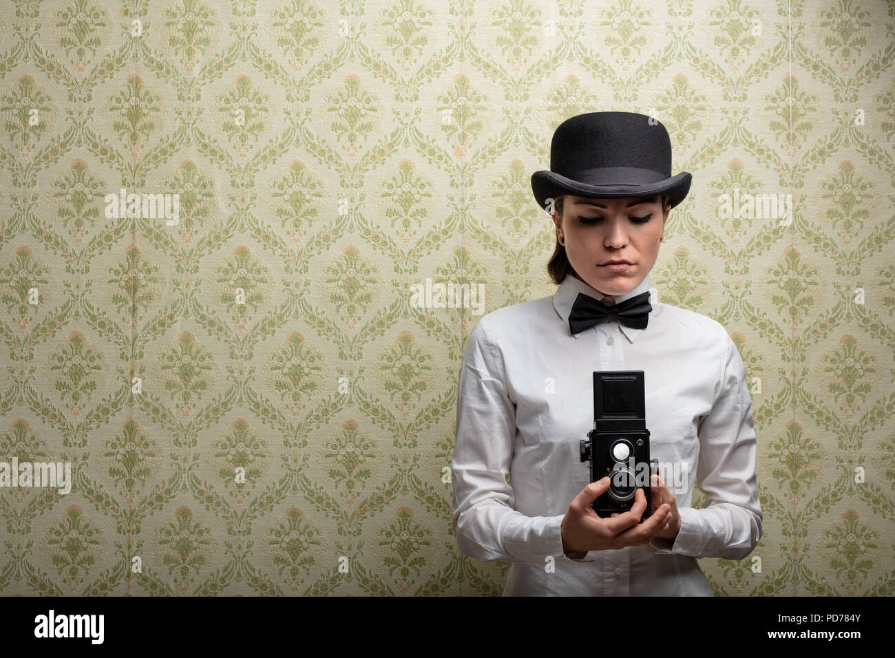 Elegant vintage woman photographer in bowler hat and bow tie holding an old camera against patterned wallpaper with copy space Stock Photo