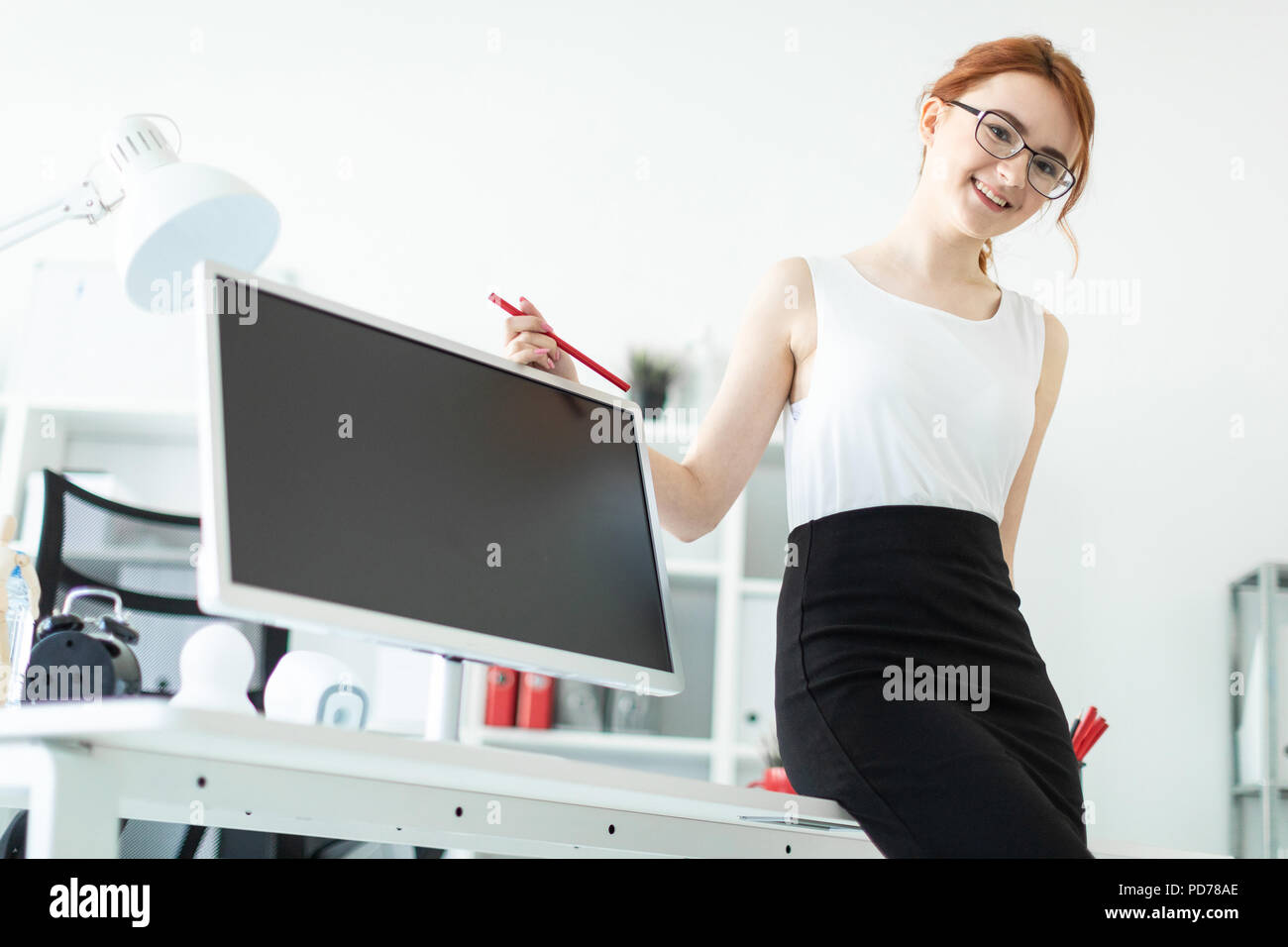 A beautiful young girl in the office sat down on the table and was holding a red pencil in her hand. Next to the girl is a monitor. Stock Photo