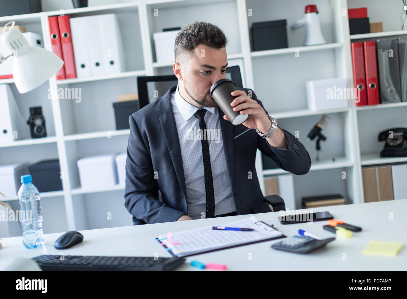 A man is sitting in the office at the table and drinking coffee from a glass. Stock Photo