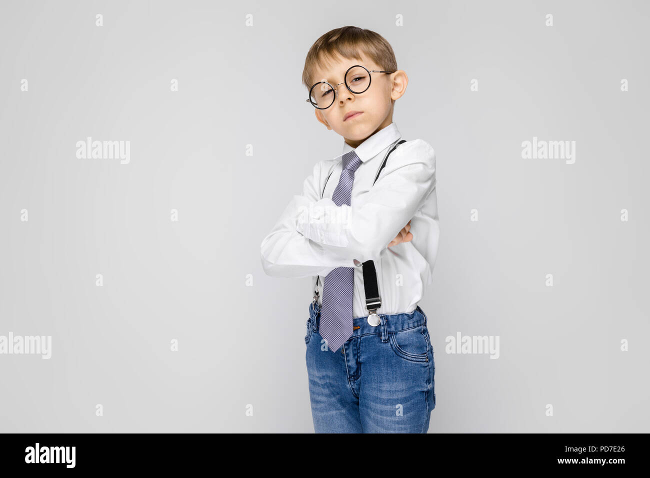 A charming boy in a white shirt, suspenders, a tie and light jeans stands  on a gray background. The boy folded his arms over his chest Stock Photo -  Alamy