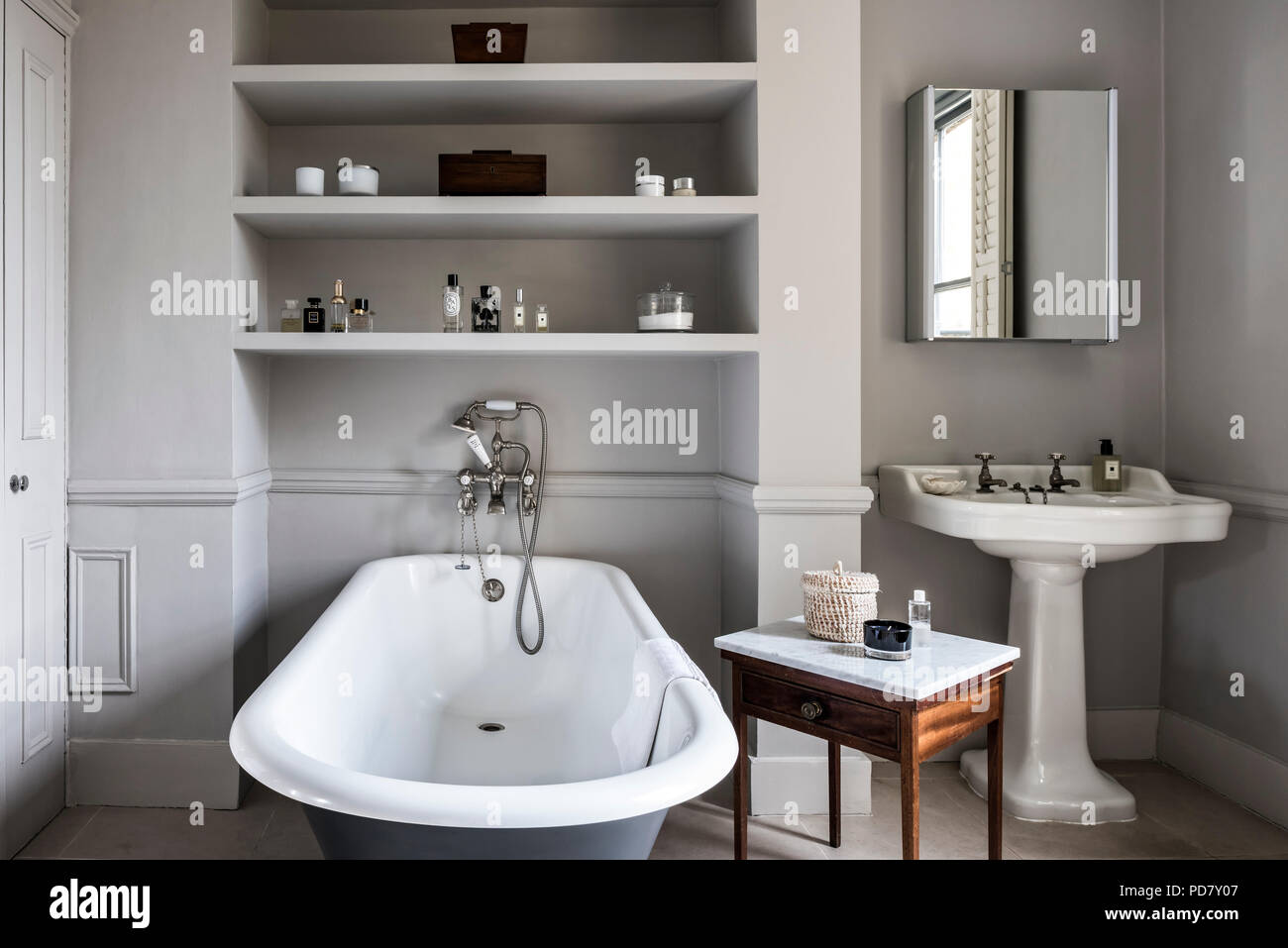 Free-standing roll top bath from Albion in bathroom with antique marble topped table and inbuilt shelving. The outside of the bath is painted in Raili Stock Photo