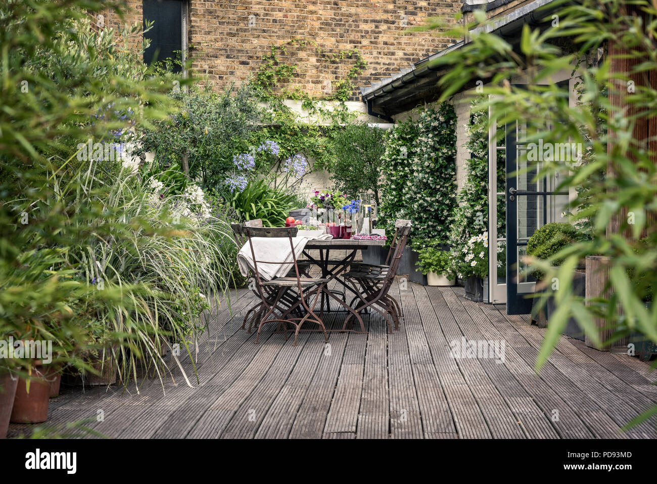 Old French bistro chairs around long wooden table on garden roof terrace with decking. Agapanthus and jasmine flourishes in the background Stock Photo