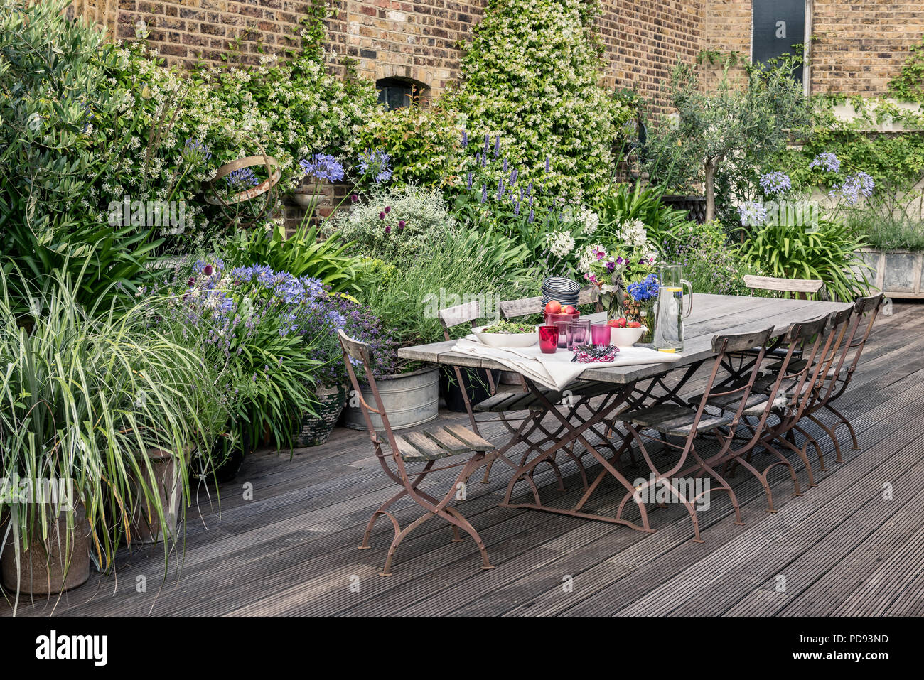 Old French bistro chairs around long wooden table on garden roof terrace with decking. Agapanthus and jasmine flourishes in the background Stock Photo