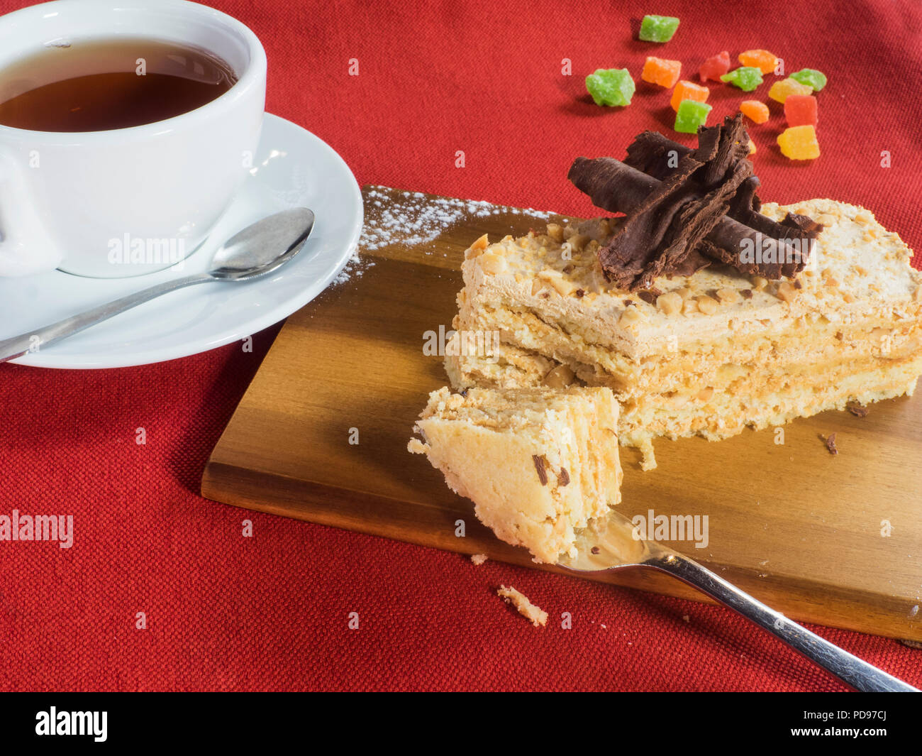 piece of shortcake cake in walnuts, decorated with crumbs and chocolate on a wooden cutting board on a red tablecloth with a cup of tea and candied fr Stock Photo
