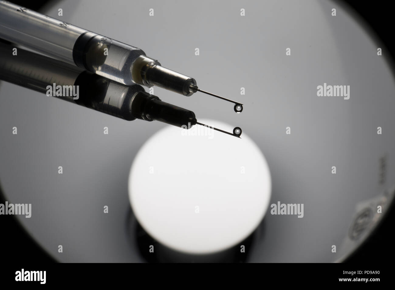 Close up of syringe with flu vaccine. Drop of liquid coming out of a needle reflecting on a shiny table. Stock Photo
