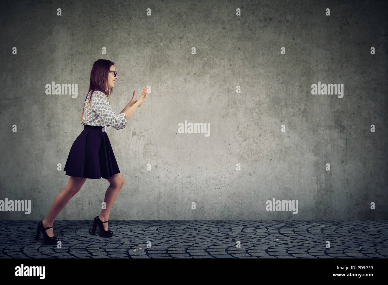 Side view of young stylish woman outstretching hands and pushing something away being strong and motivated while standing on gray wall Stock Photo