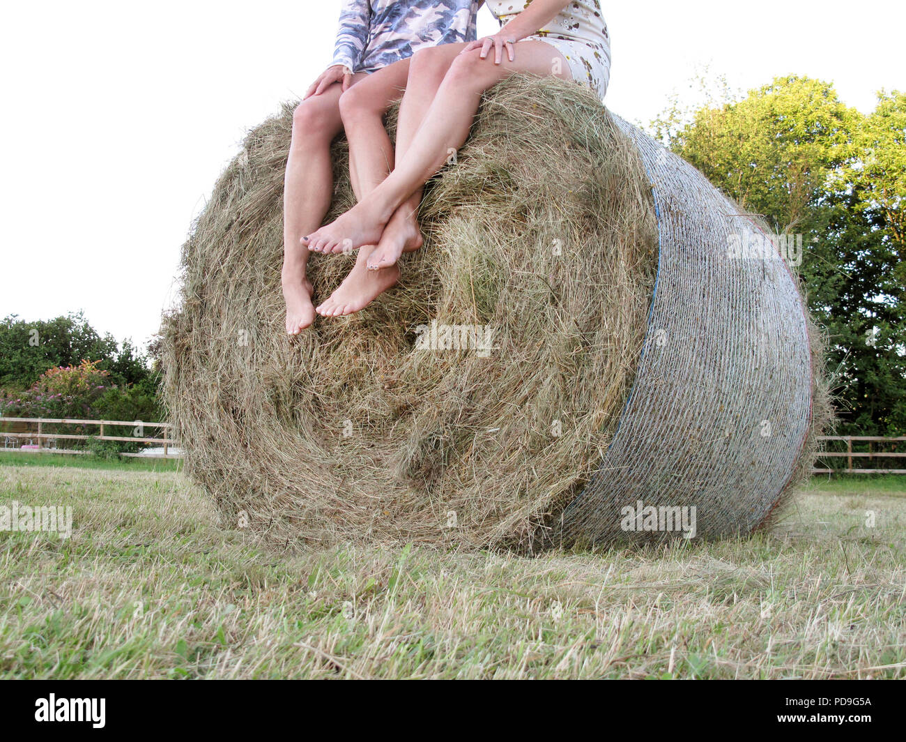 two women sitting on a roll of hay with legs around each other Stock Photo