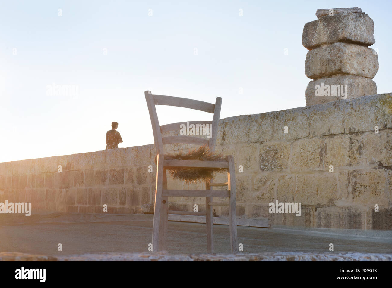 silhouette of a lonely woman sitting on a stone wall with a derelict chair in the foreground Stock Photo