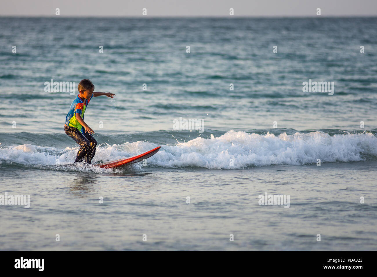 A young local boy practices his surfing skills on a small wave on a surf board donated by visiting westerns at the Tip of Borneo, Borneo, Malaysia. Stock Photo