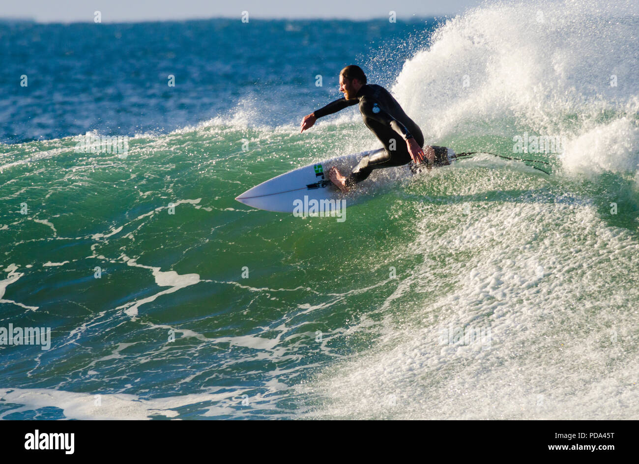 A surfer skillfully carves across a beautiful green wave, wearing a black wetsuit and riding a white surfboard Stock Photo