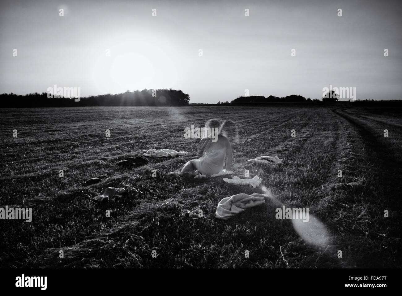 Young woman sitting in the middle of a meadow during sunshine with scattered cloth. Long exposure shot girl with two heads symbolic psychic disorder. Stock Photo