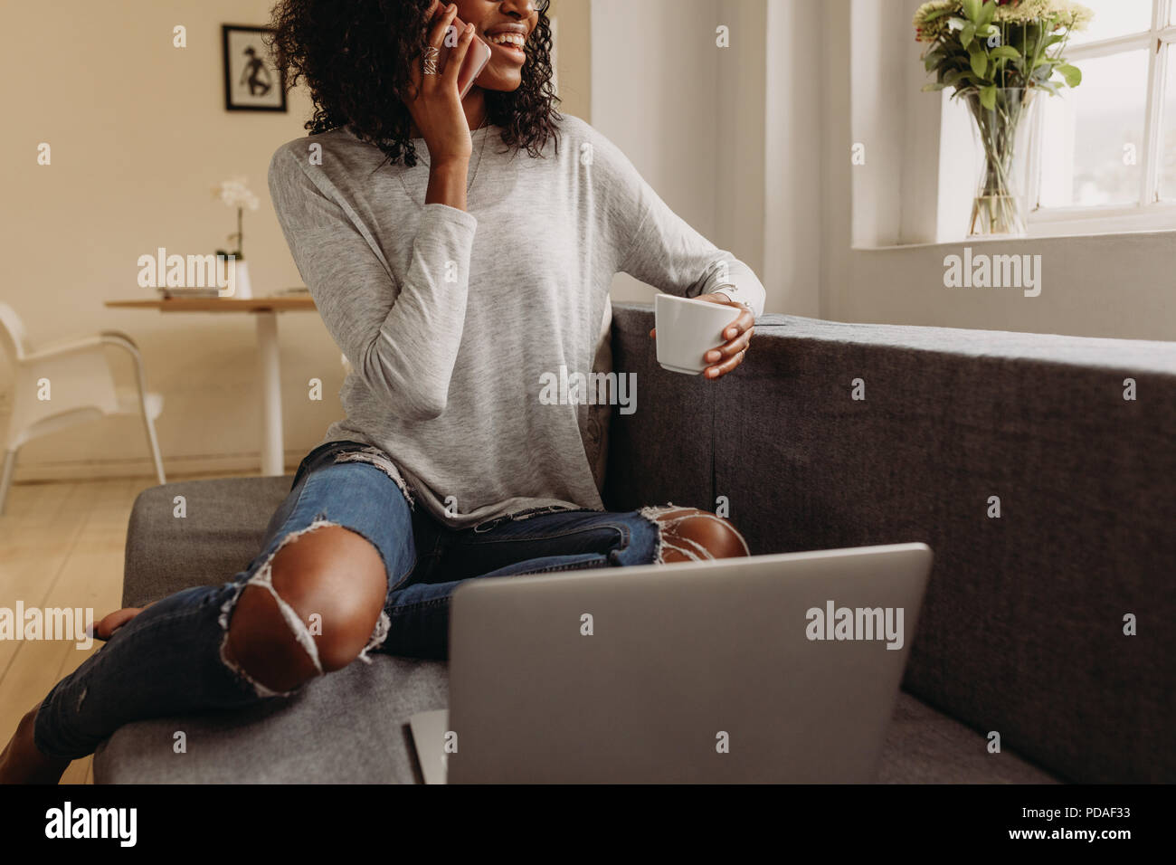 Businesswoman sitting on sofa at home and talking over mobile phone while working on laptop. Smiling woman in fashionable torn jeans holding a coffee  Stock Photo