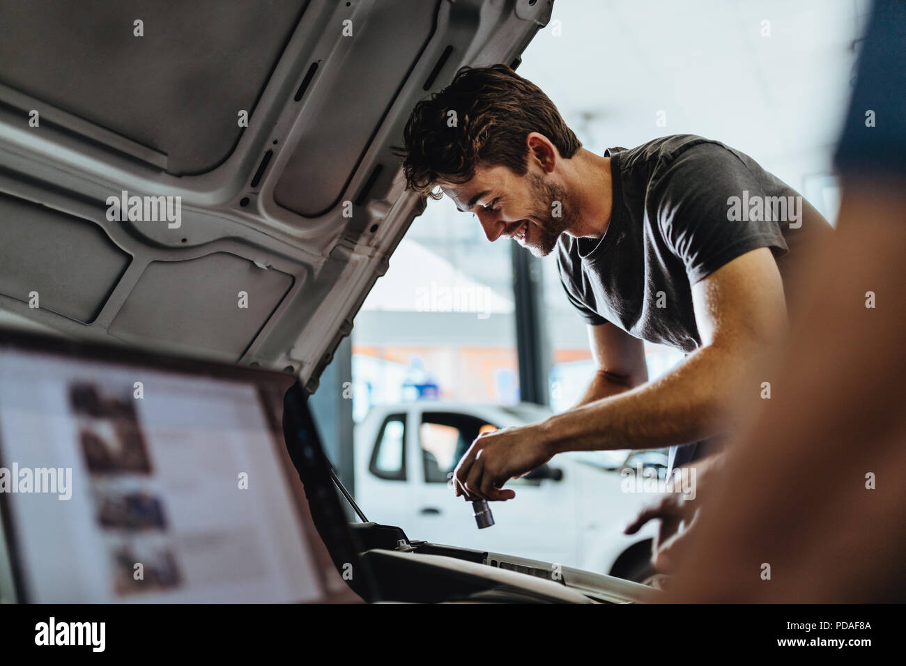Smiling young man repairing the car in garage with coworker using laptop in front. Mechanic working under the hood of a vehicle. Stock Photo