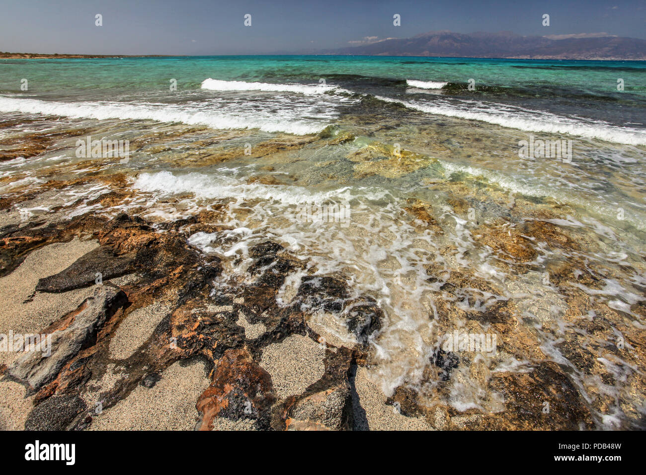Sand on large rocks, crystal clear turquoise sea with white waves and land in background. Chrissi , Greece Stock Photo