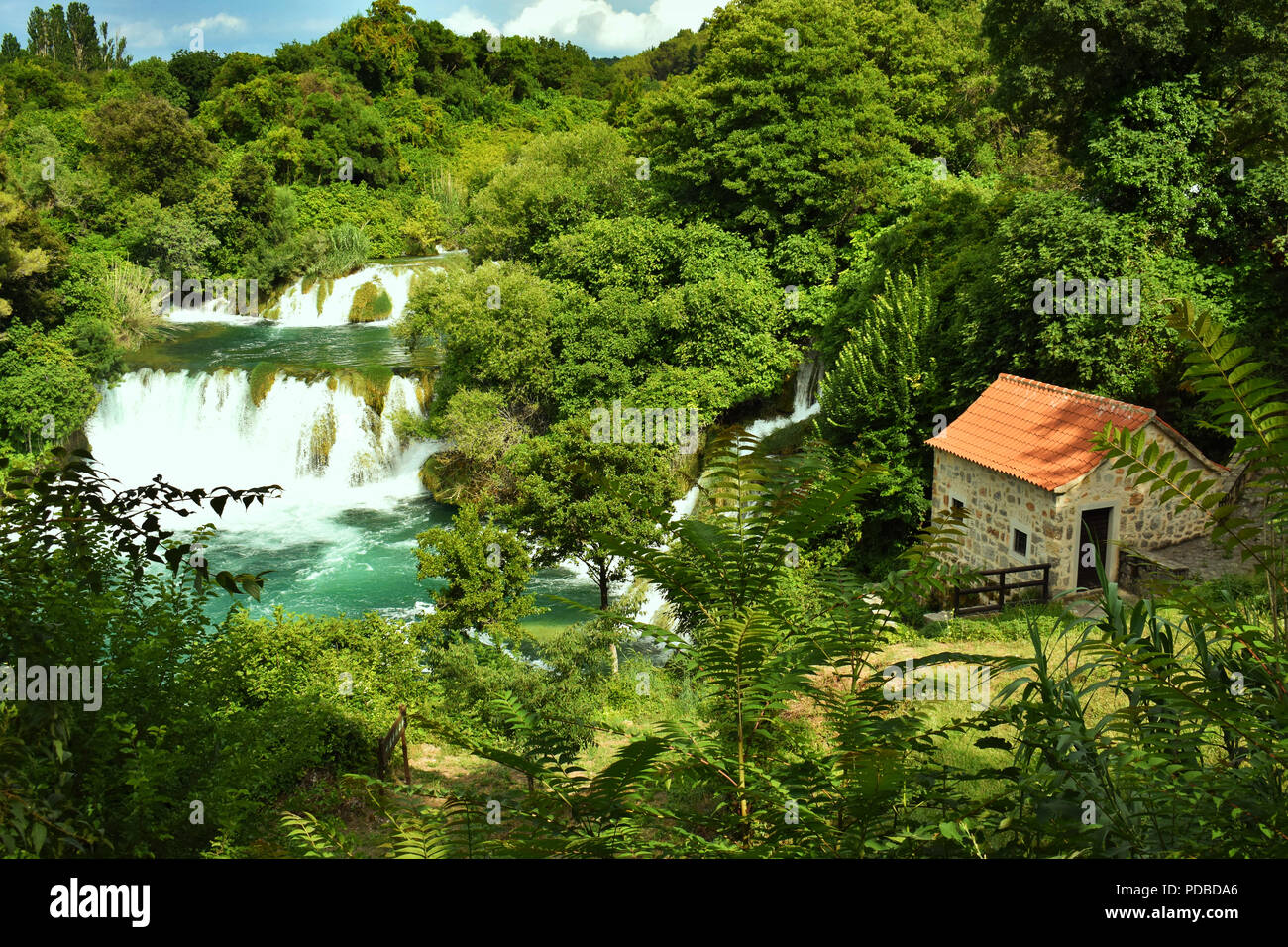 Small House Next to Beautiful Clear Turquoise Waterfalls and Surrounded by Green Trees in the Forrest With Blue Sky and Clouds Stock Photo