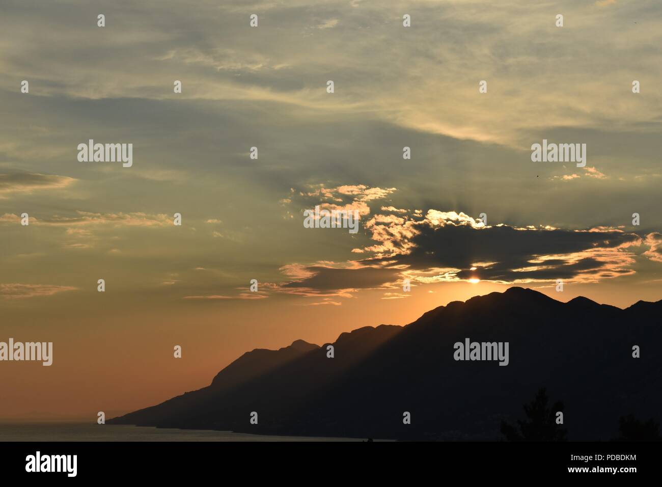 Bright Orange Sunset Silhouetting the Mountains of the Dalmatian Coast Croatia with Sunlight Beaming through the Clouds Stock Photo