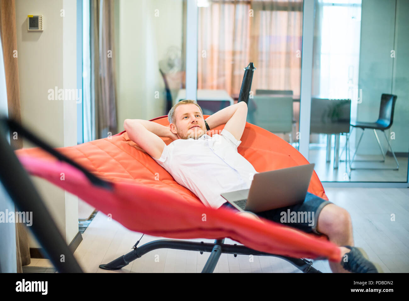 Young man with laptop holding hands behind head, relaxing and dreaming while lying on a hammock in office space. Taking time for a minute break. Unpro Stock Photo