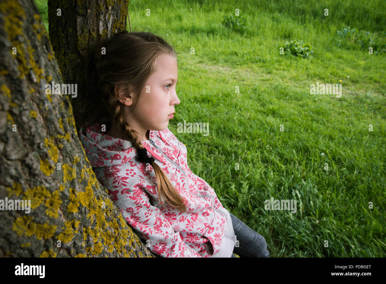 child girl leaning on tree trunk in pensive mood outdoors Stock Photo