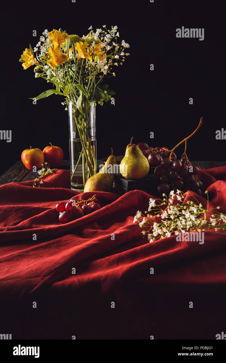 still life with fruits and flowers in vase on red drapery on black ...