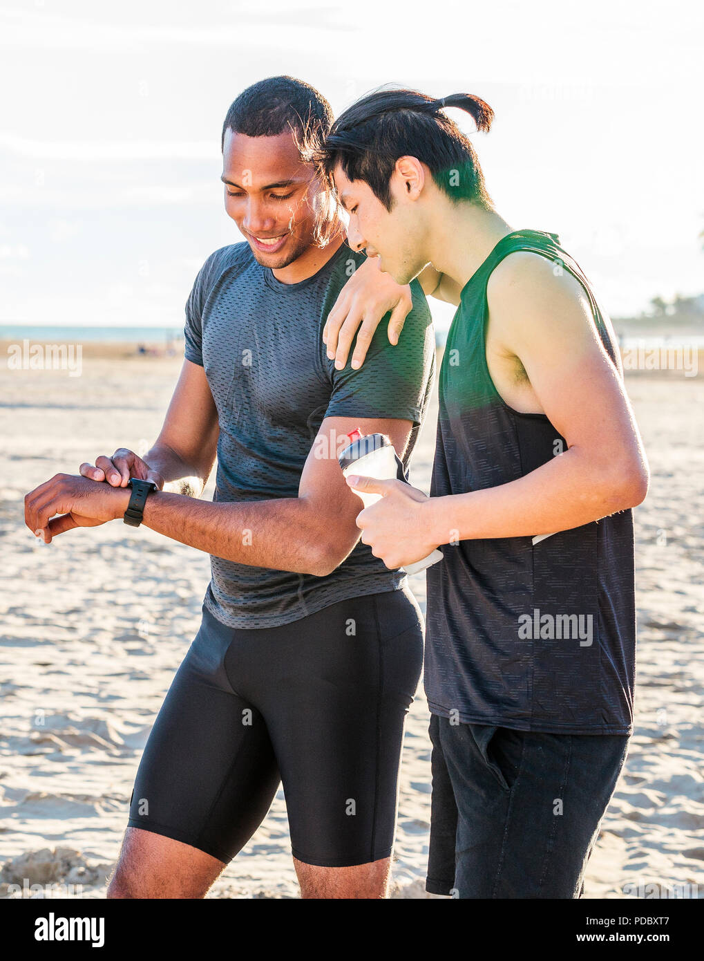 Male runners resting, checking smart watch fitness tracker on sunny beach Stock Photo