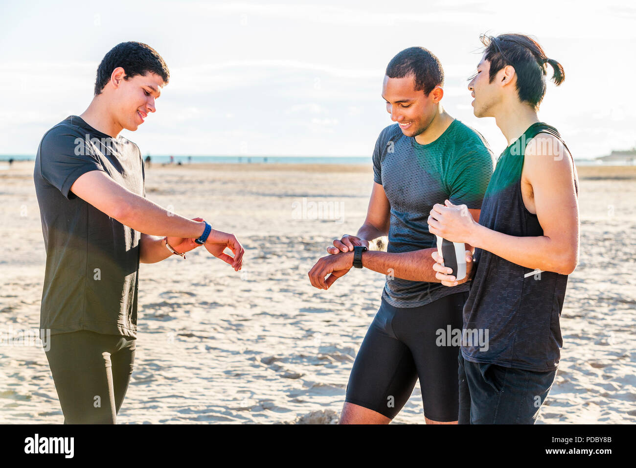 Male runners checking smart watch fitness trackers on sunny beach Stock Photo