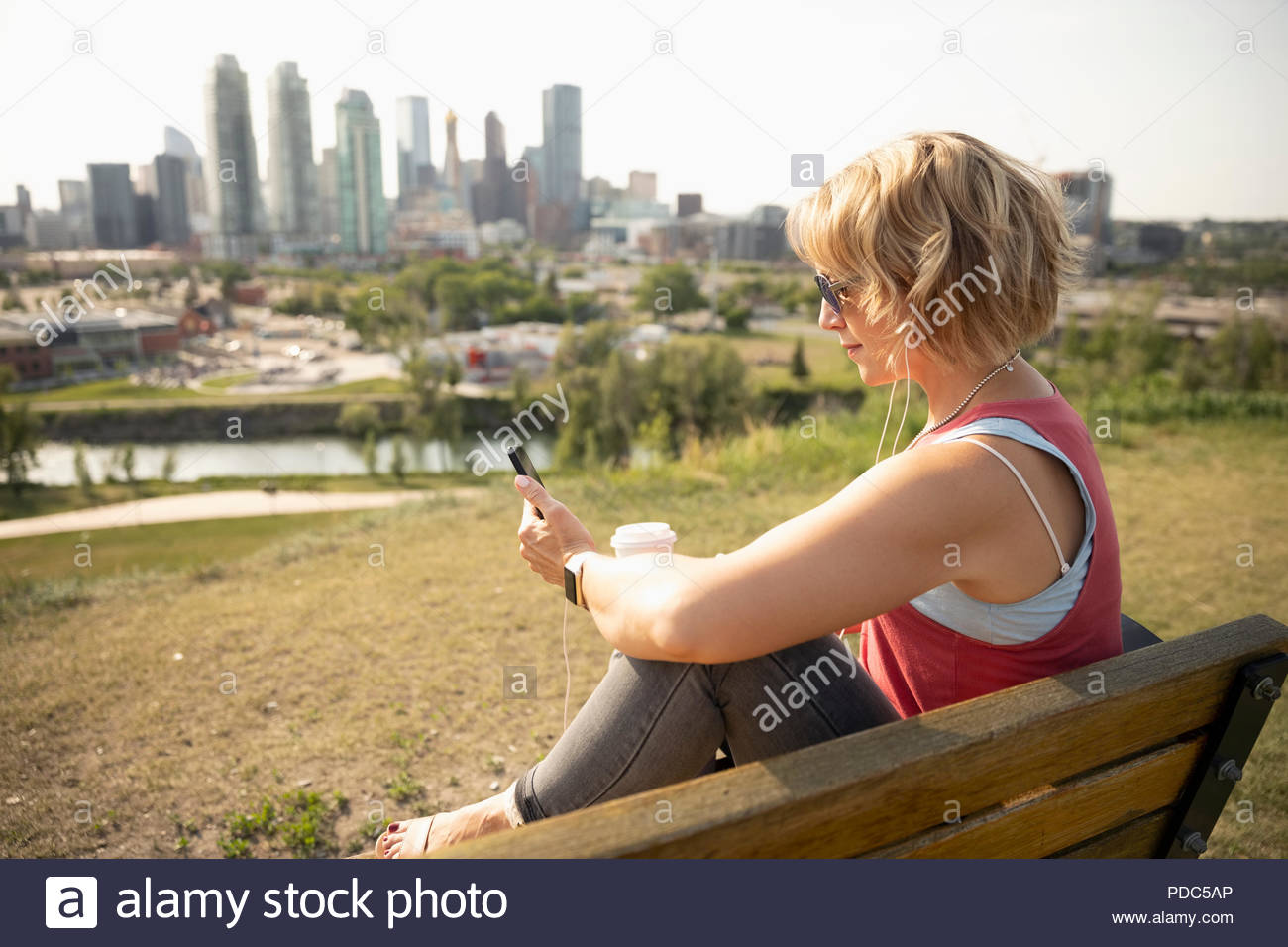 Woman with coffee listening to music with headphones and mp3 player on sunny urban park bench Stock Photo