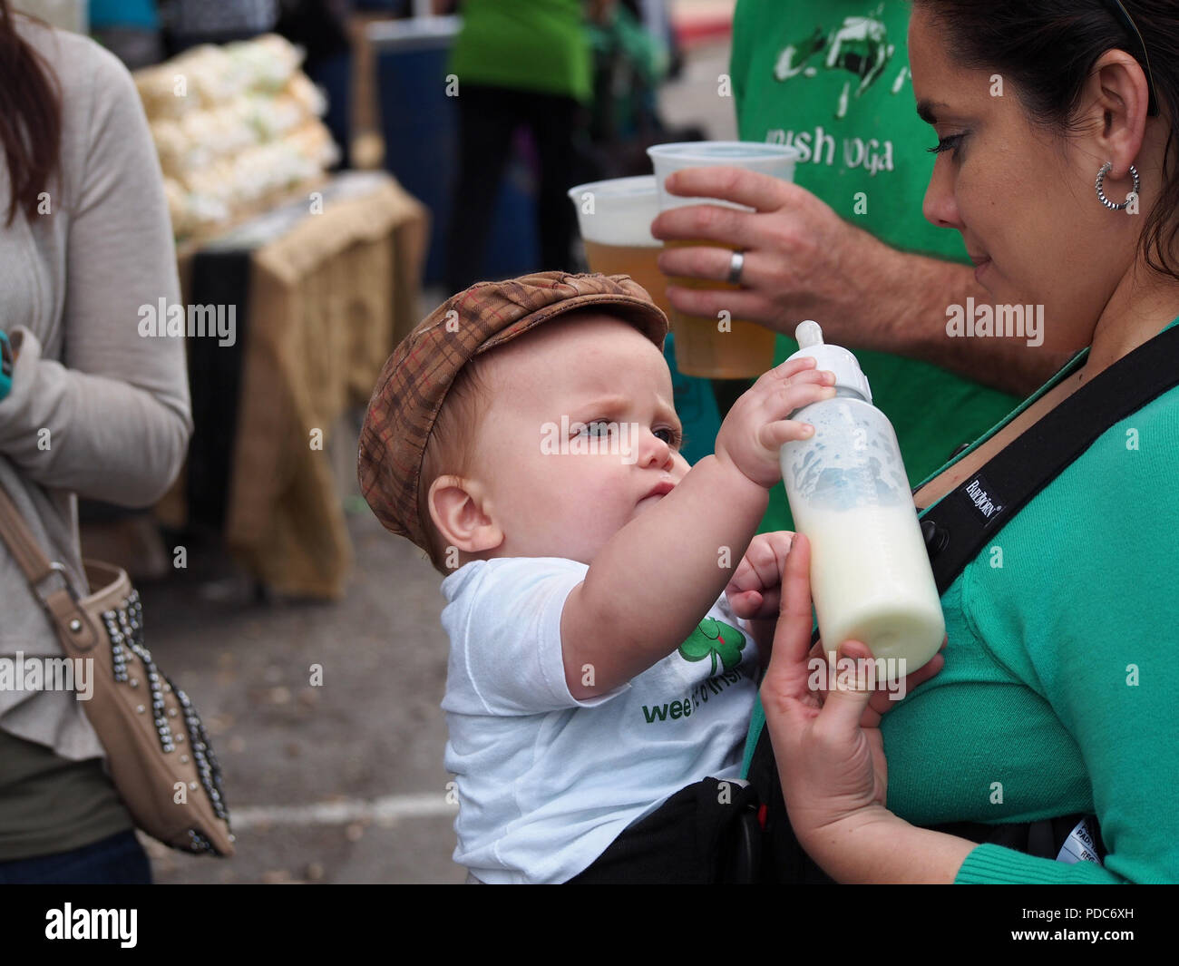 A baby boy in a tweed cap reaches for his bottle under his Mother's loving gaze. St. Patrick's Day Block Festival in Corpus Christi, Texas USA. Stock Photo