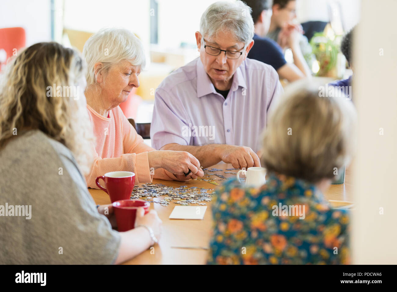 Senior friends assembling jigsaw puzzle and drinking tea at table in community center Stock Photo