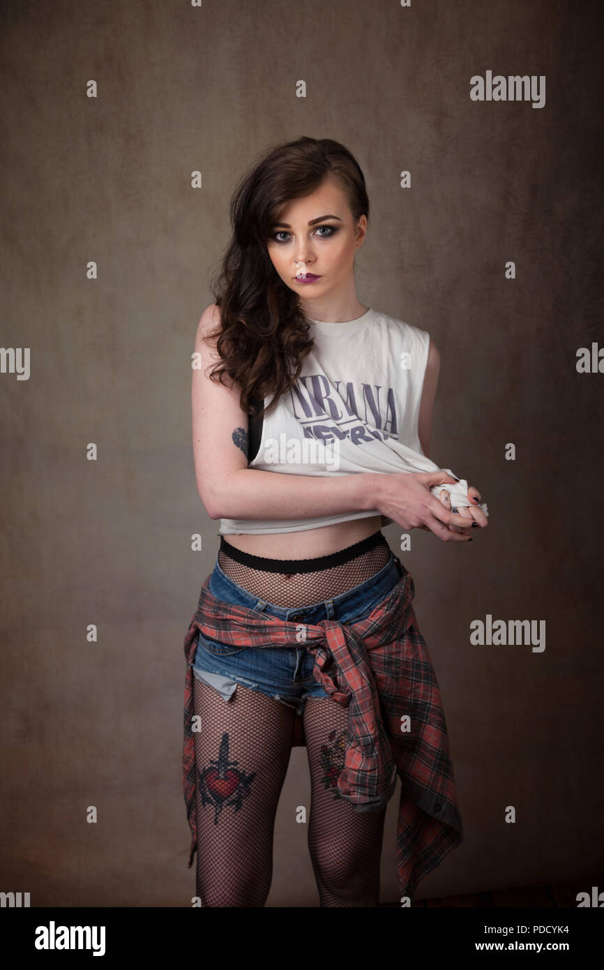 An alternative looking girl standing in a studio wearing a band vest top and fishnet tights which show above her denim shorts. Stock Photo
