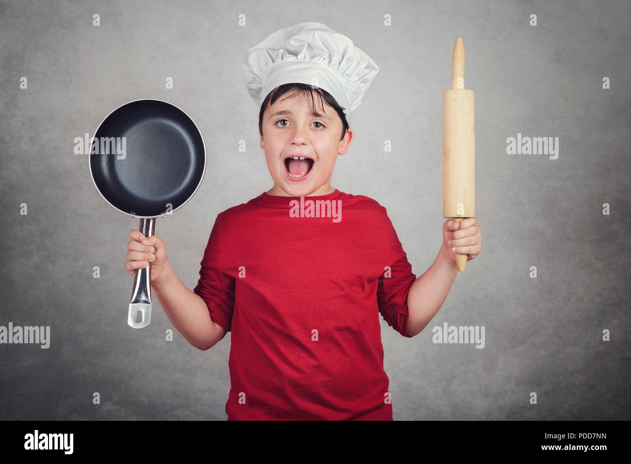 Happy little boy in chef uniform with a rolling pin and frying pan on gray background Stock Photo