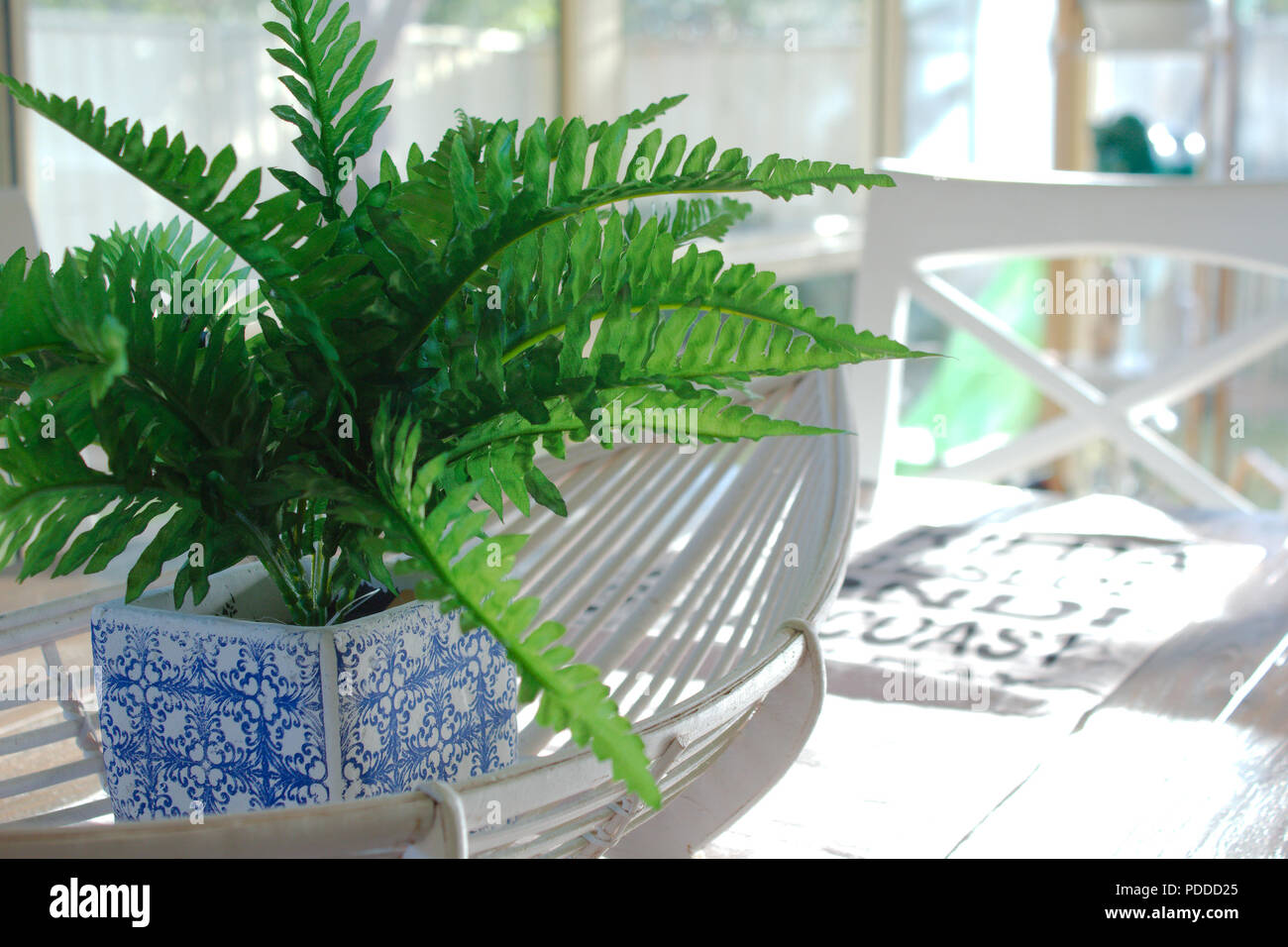 A fake green fern plant in a blue & white plant pot sits in a white cane oval basket. The table decor sits in the centre of a wooden dining table. Stock Photo