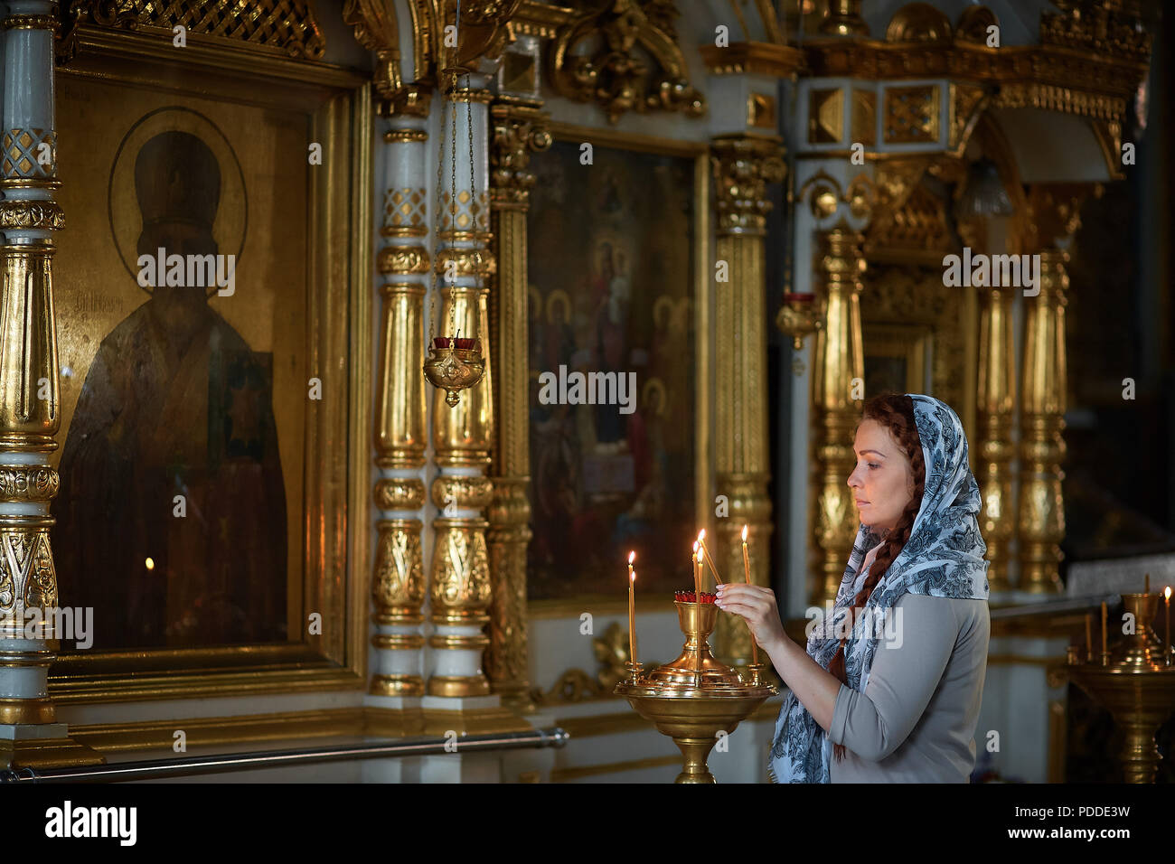 Russian beautiful Caucasian woman with red hair and a scarf on her head is in the Orthodox Church, lights a candle and prays in front of the icon Stock Photo
