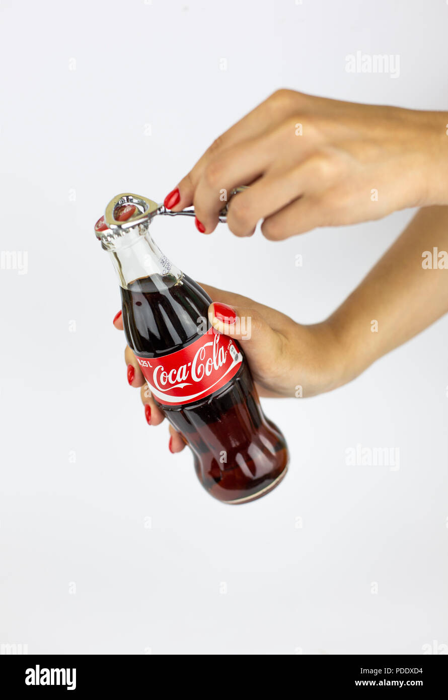 Atlanta, Georgia, USA - July 22, 2018: hand red nails holding historical embossed glass coca-cola contour classic bottle from Spain on white background opening with metal coca-cola brand opener