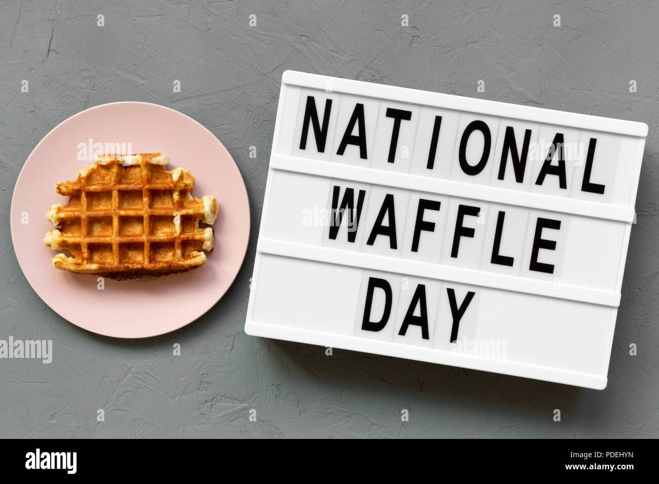 'National Waffle Day' word on lightbox, traditional belgian waffle on pink plate over concrete