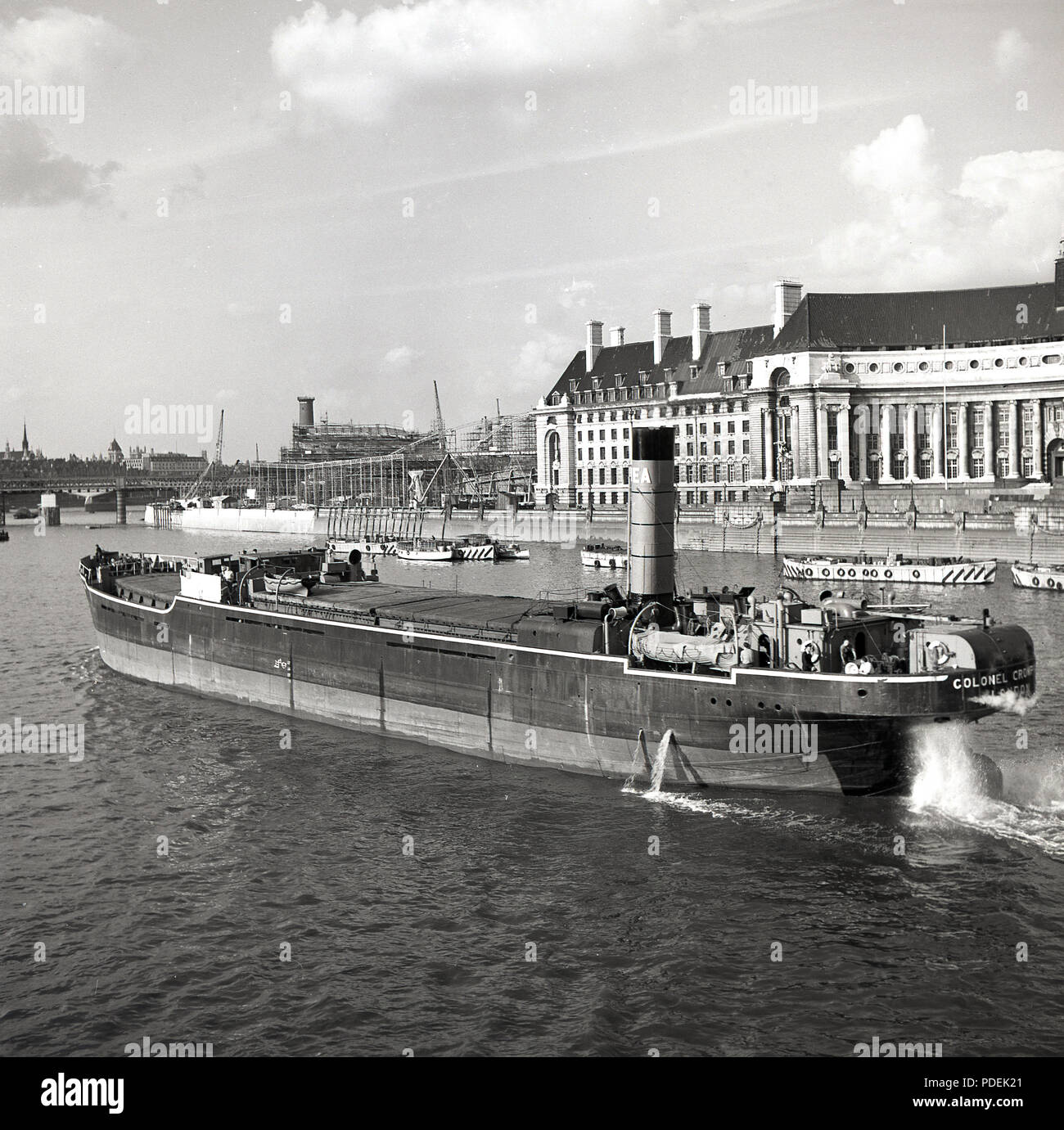 1950s, a large cargo steam barge on the River Thames carrying freight goes past County Hall, home of the LCC, on the South Bank, London, England, UK. In this era, the river was very much a working one. On the left of County Hall can be seen the construction of the Royal Festtival Hall. Stock Photo