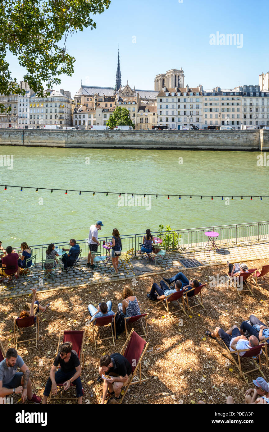 People resting on deck chairs along the Seine during Paris-Plage summer event in Paris, France, with Notre-Dame de paris cathedral in the background. Stock Photo