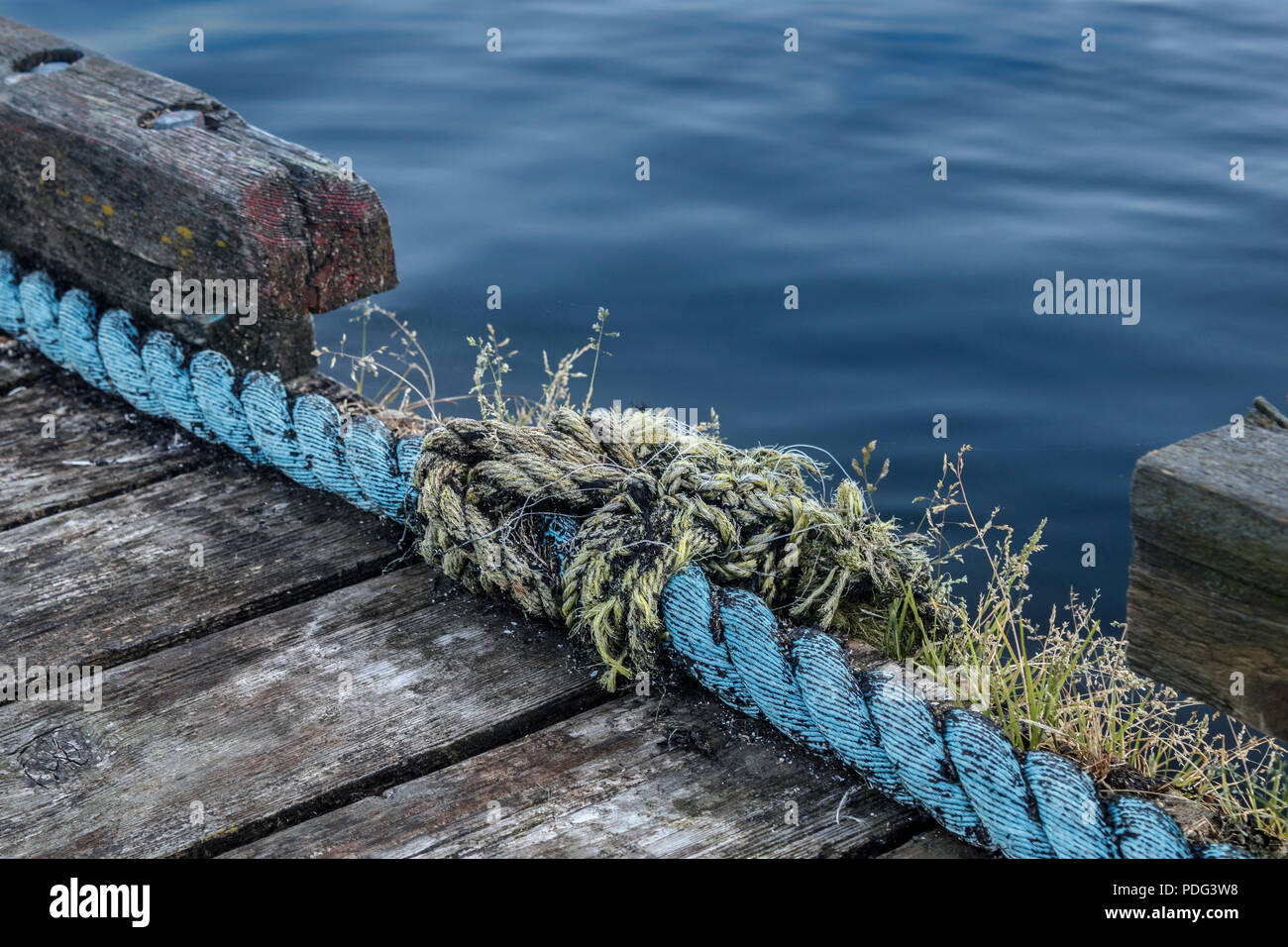 The smooth water surface contrasts with the rough textures of a weathered old wooden dock and the once-strong but now fraying ropes strung along it. Stock Photo