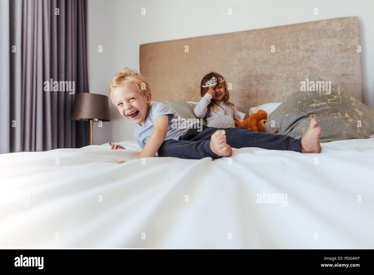 Happy kids playing in bedroom. Little boy smiling with girl at the back in crown on the bed. Stock Photo
