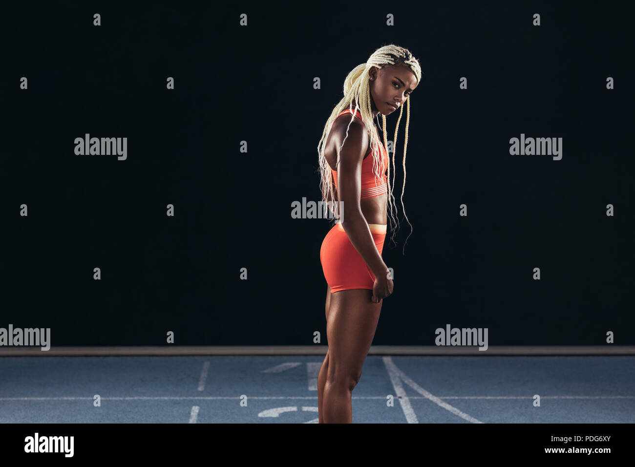 Female sprinter standing at the start line on a running track on a black background. Side view of a female runner standing at the start line on an all Stock Photo