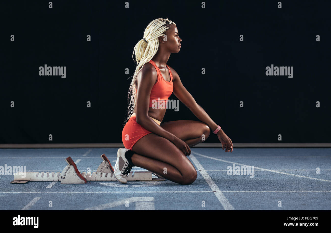 Female sprinter sitting at the start line near the starting block on a running track. Side view of female runner sitting at the start line on running Stock Photo