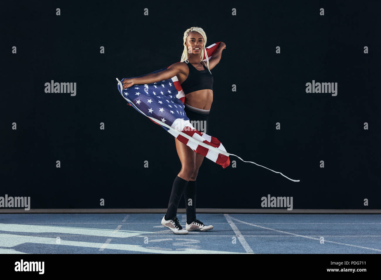 Female athlete walking on running track celebrating victory holding american flag. Sprinter walking on running track carrying the US flag behind her. Stock Photo