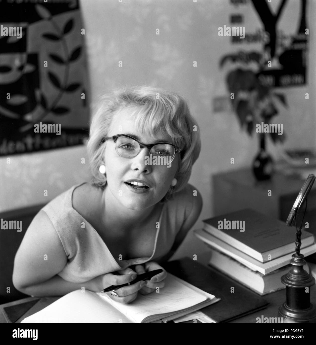1950s sunglasses. A young woman is sitting at a desk looking into the camera wearing a pair of glasses with  typical 1950s bows. Sweden 1957. Ref 3509    Lena Granhagen. Swedish actress born 1938. Stock Photo
