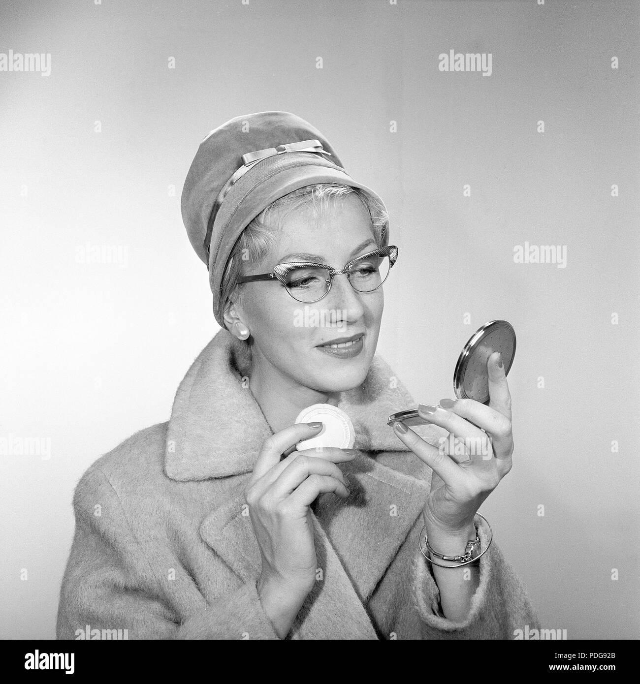 1950s makeup. A young woman is looking at herself in her pocket mirror and improves on her makeup. She is wearing a fashionable hat, typical 50s glasses and a coat. 1950s Stock Photo