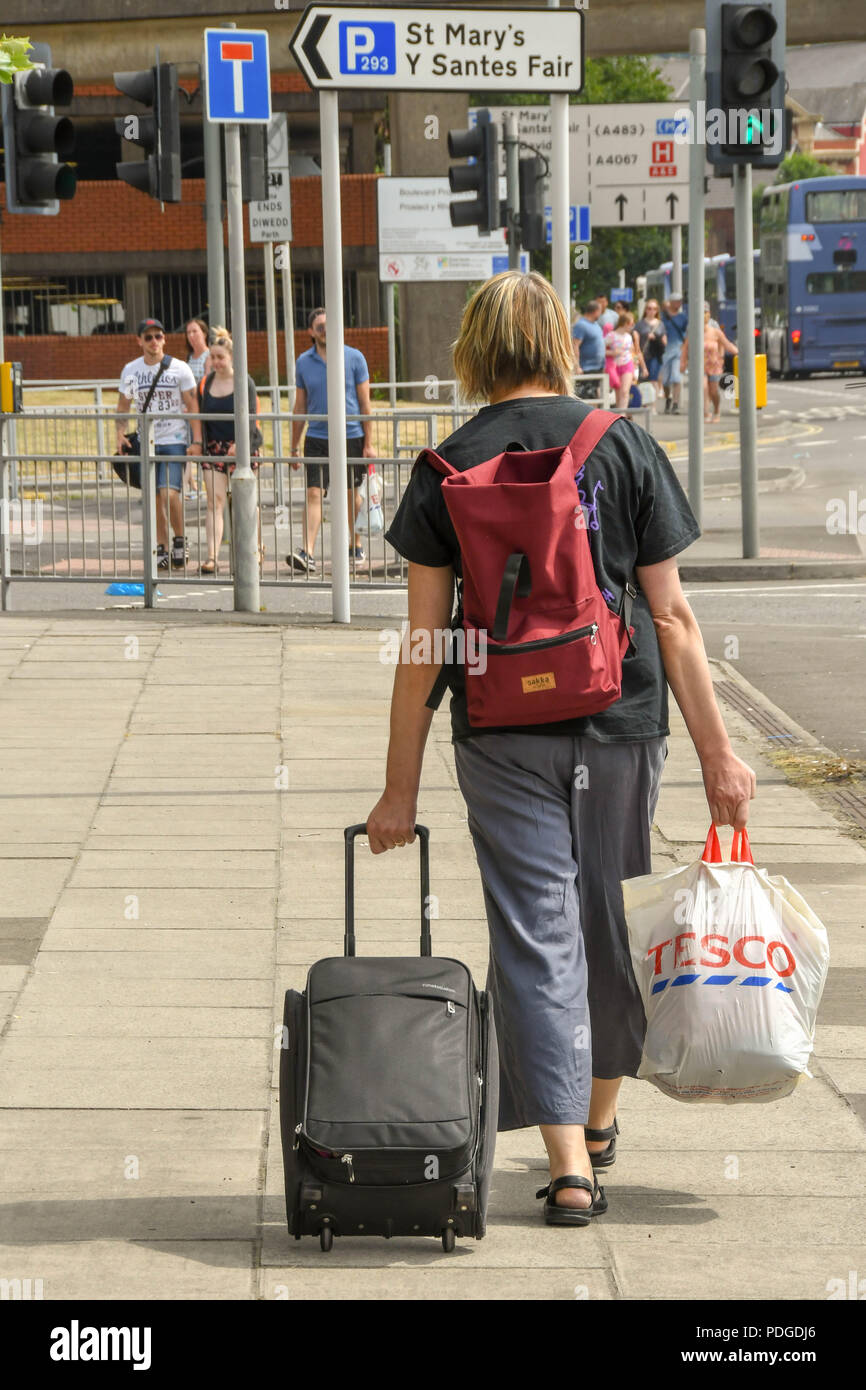 Person pulling a wheeled suitcase and carrying a plastic bag walking along a street in Swansea, Wales Stock Photo